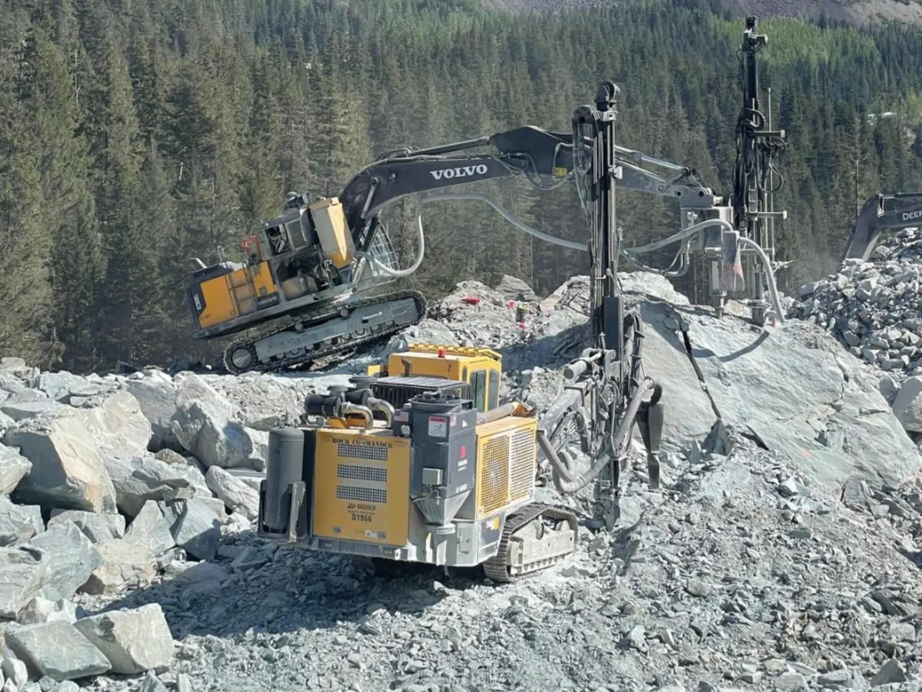 McCaw's drill rigs and an excavator working freshly blasted rock on a forested job site
