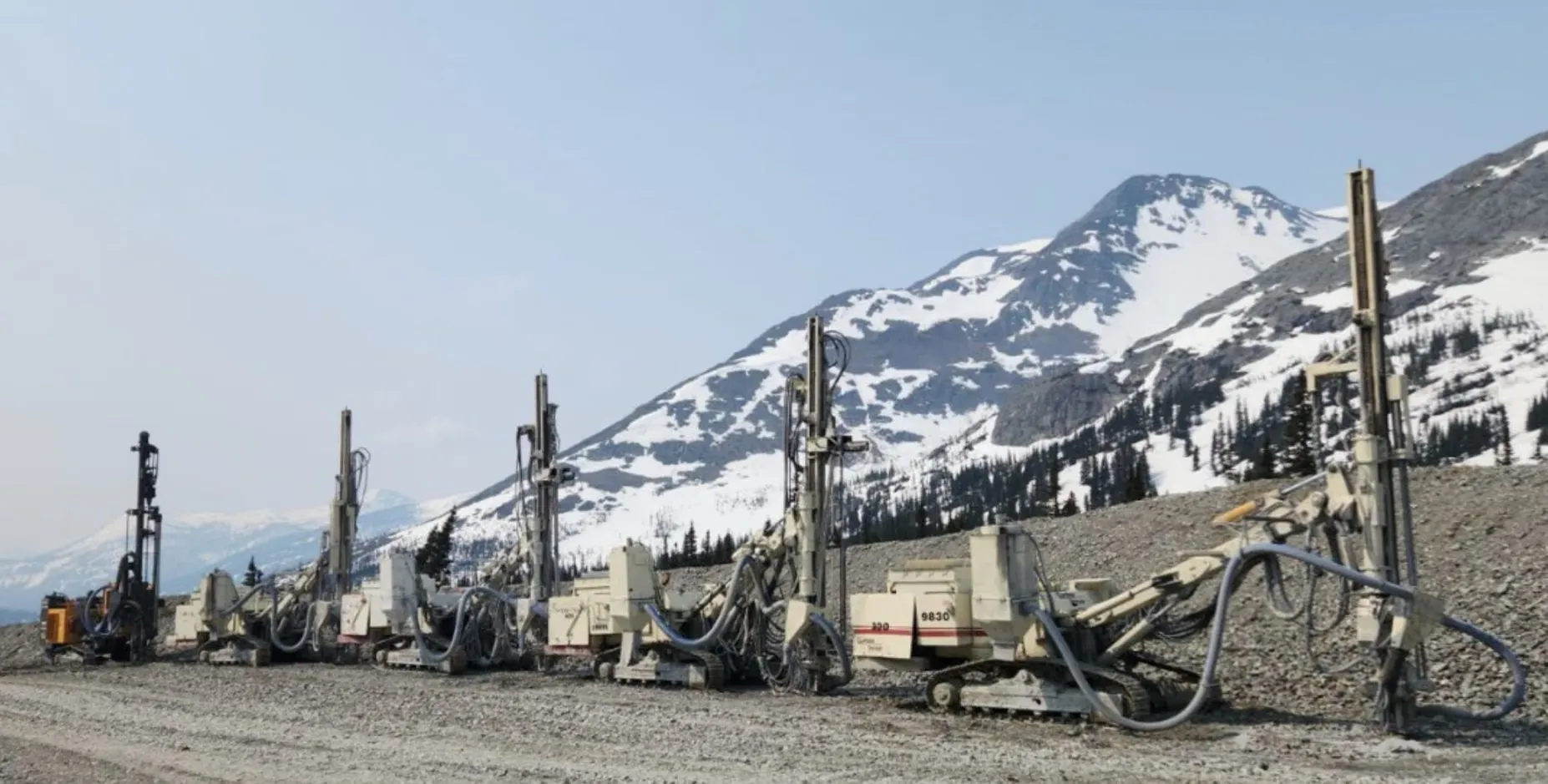 A lineup of McCaw's drill rigs staged on a gravel pad below a snow-covered BC mountain